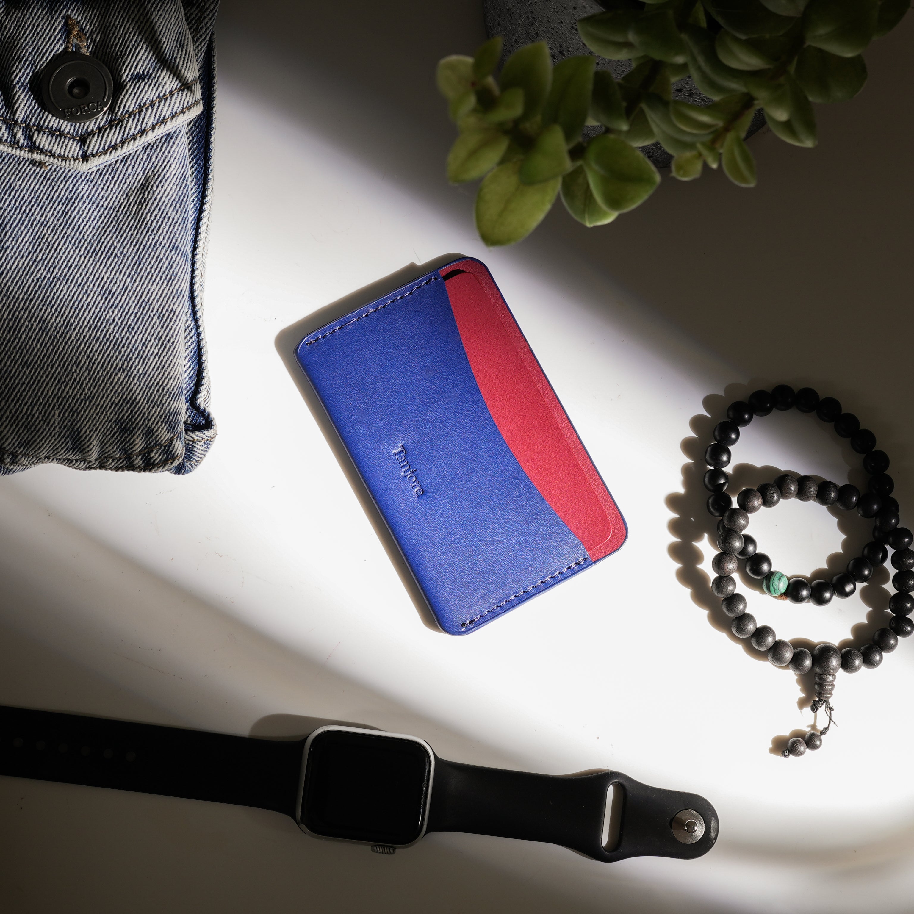 Flatlay of a Navy and Plum Tanjore cardholder on a white desk next to a watch and denim jacket. The Full Grain Nappa Leather texture is highlighted in natural light, backed by a 3 year warranty.