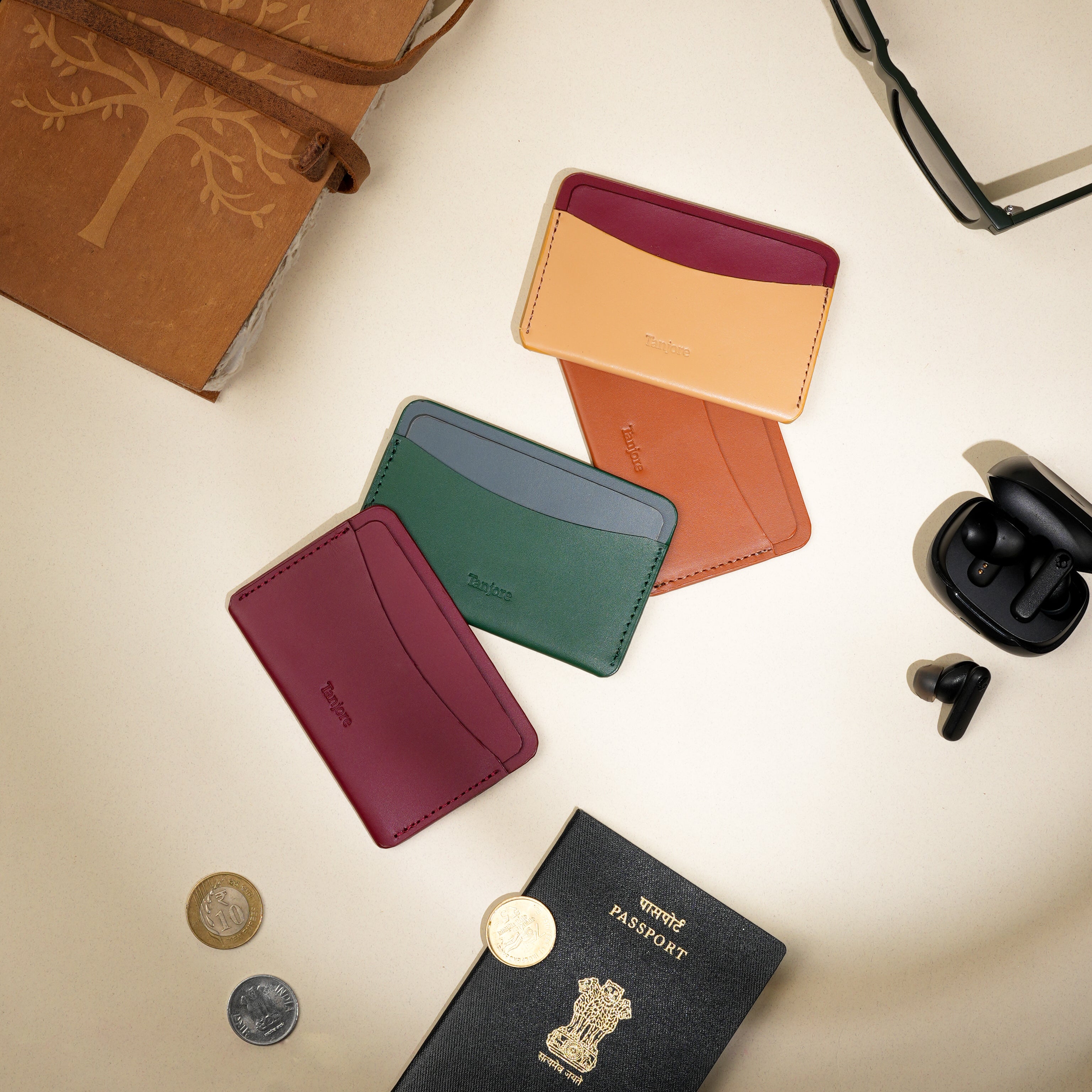 Flatlay featuring the Tanjore Clay and Plum wallet alongside green and red variations, a passport, and earbuds. Luxury Full Grain Nappa Leather travel essentials with a 3 year warranty.