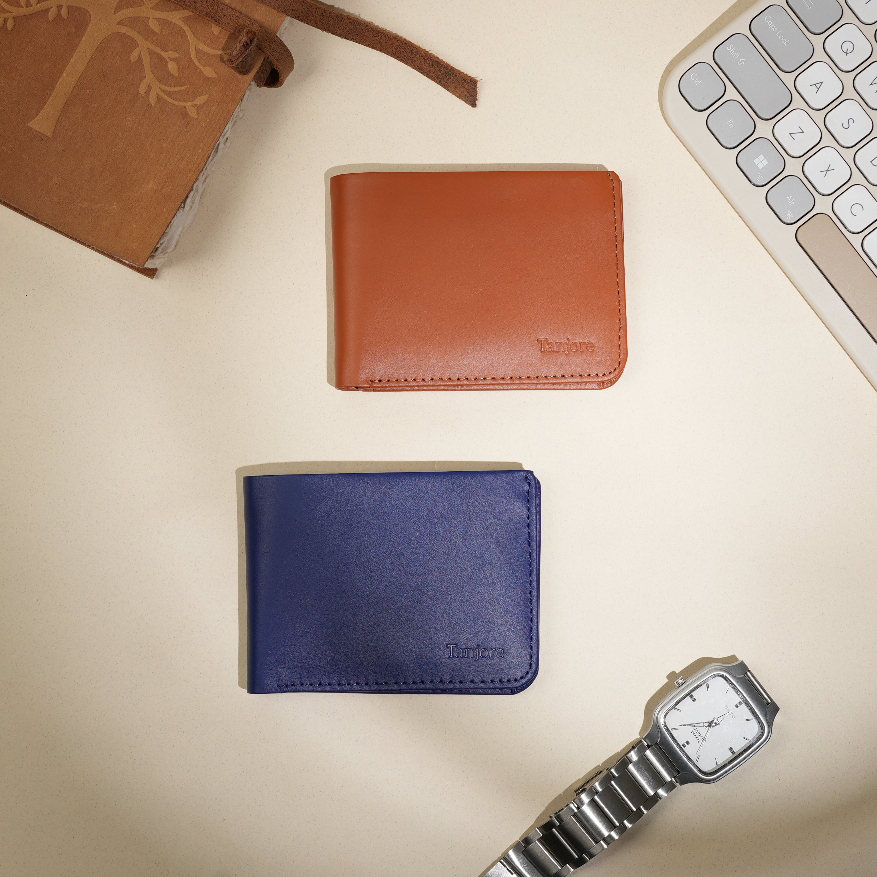 Top-down view of Tanjore Modern Classic 2.0 wallets in Tan and Navy, styled on a desk with a keyboard and watch. Premium Full Grain Nappa Leather bifolds perfect for professional settings.