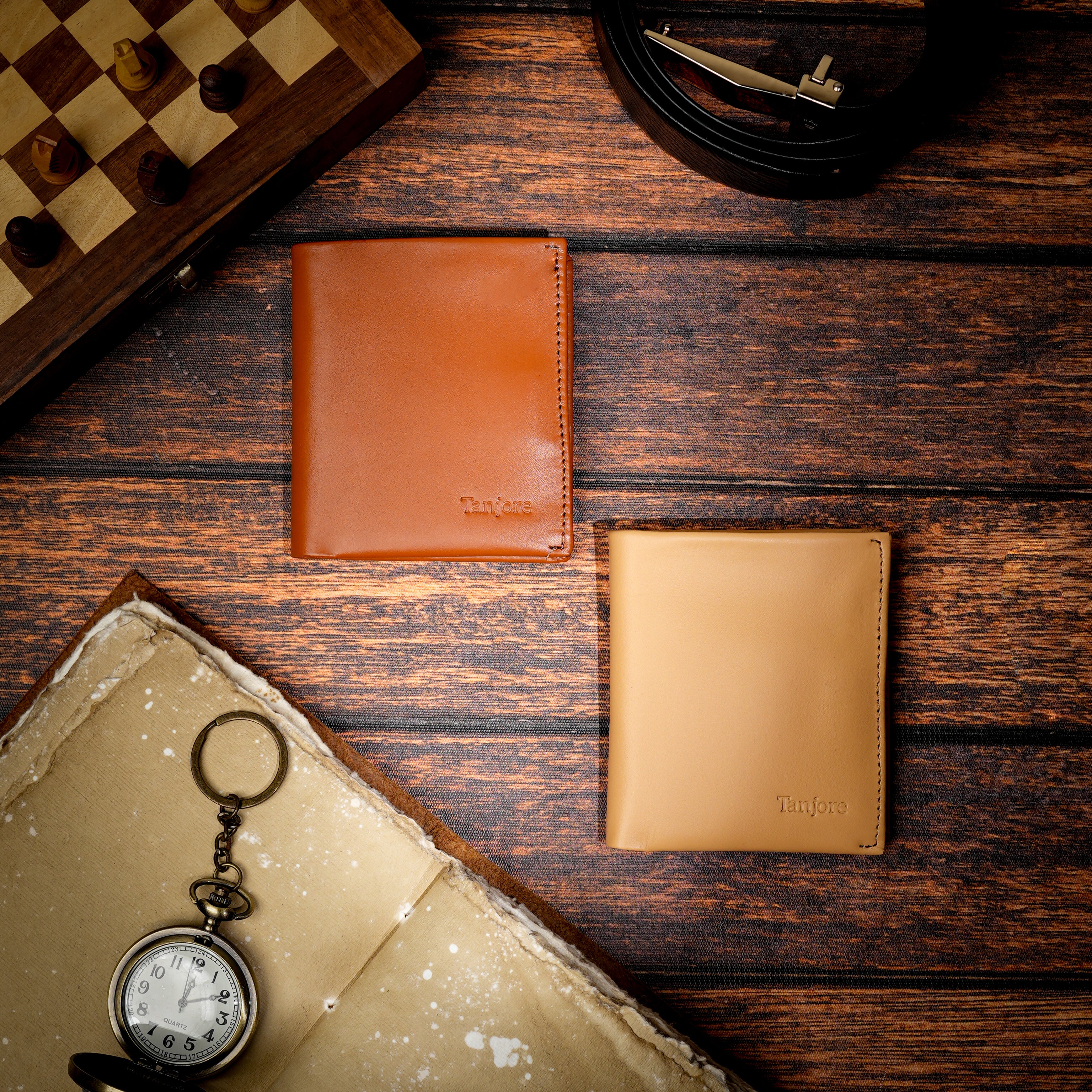 Two Tanjore Reserve Sleeves in Tan and Brown displayed on a wooden surface with a vintage pocket watch and chess board. Timeless Full Grain Nappa Leather accessories.